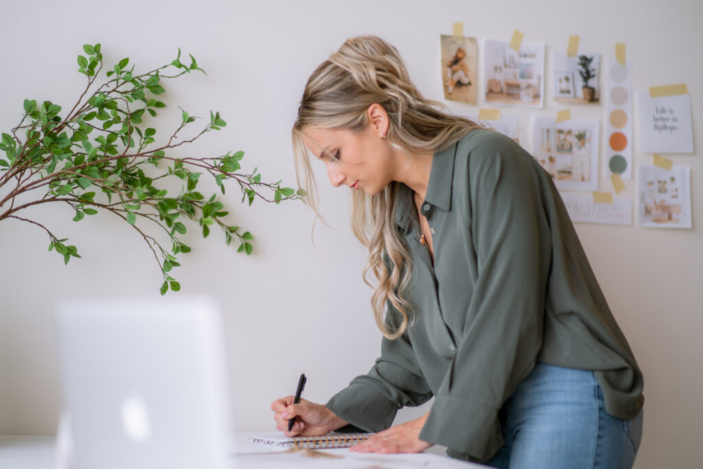 Writing a brand strategy checklist in a notebook beside a laptop and inspiration board.
