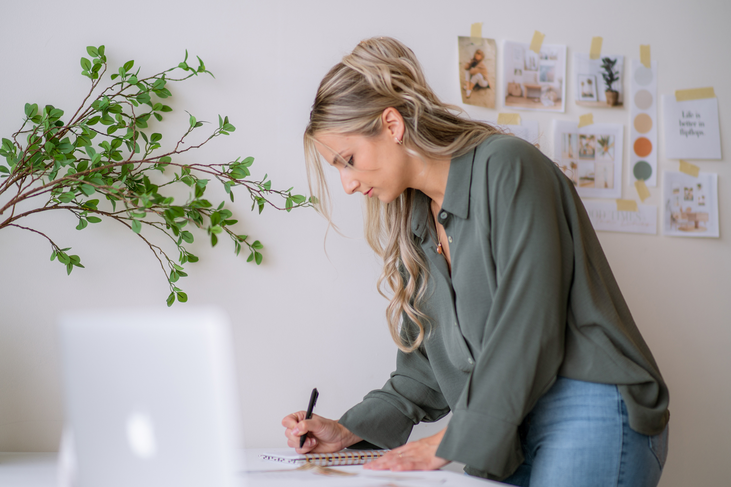 Writing a brand strategy checklist in a notebook beside a laptop and inspiration board.